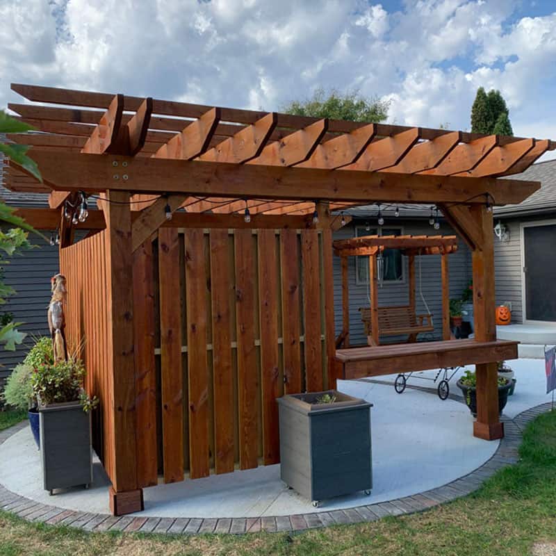 A wooden pergola with a slatted roof and a hanging porch swing underneath stands on a circular concrete patio. There are two planters on either side of the entrance. The sky is partly cloudy, and the background features parts of neighboring houses.