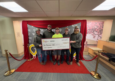 Six men stand together on a red carpet, smiling and donating a large ceremonial check for $2,000 to the Children's Miracle Network
