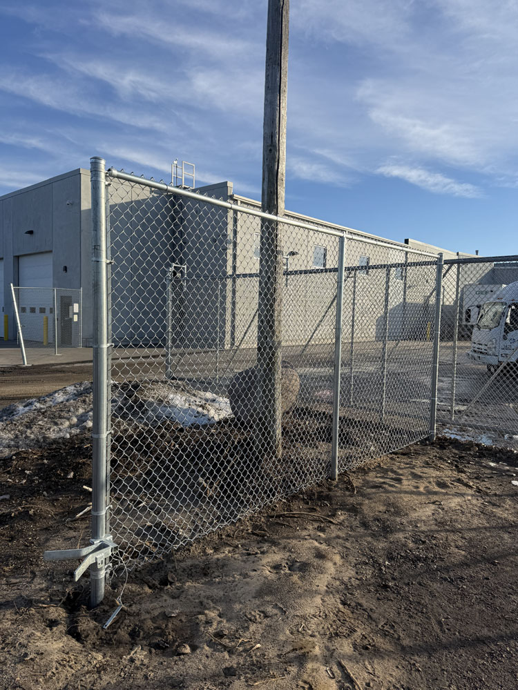 A chain-link fence is built around a utility pole, with the pole passing through the fence. There is snow on the ground and an industrial building in the background under a blue sky.