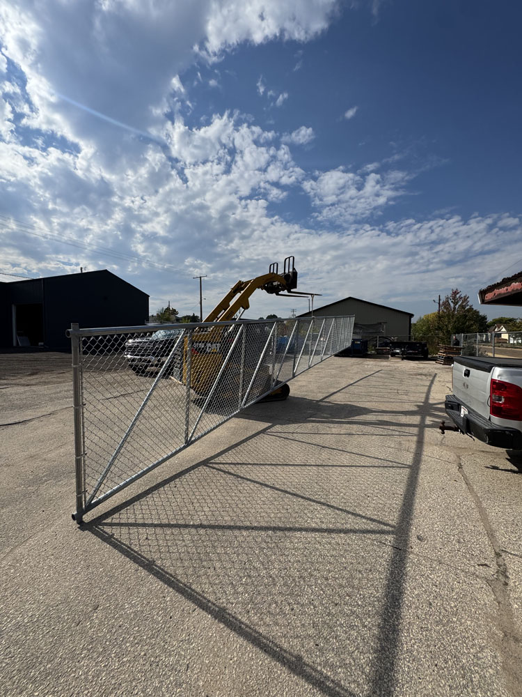 A construction vehicle lifts and installs a tall chain-link fence on a sunny day in a parking lot, with blue buildings and a white truck nearby under a partly cloudy sky.