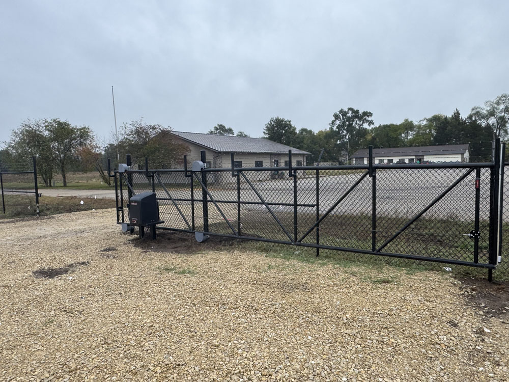 A black metal chain-link gate with an automatic opener stands on a gravel driveway, leading to a fenced area with trees and a building in the background under a cloudy sky.