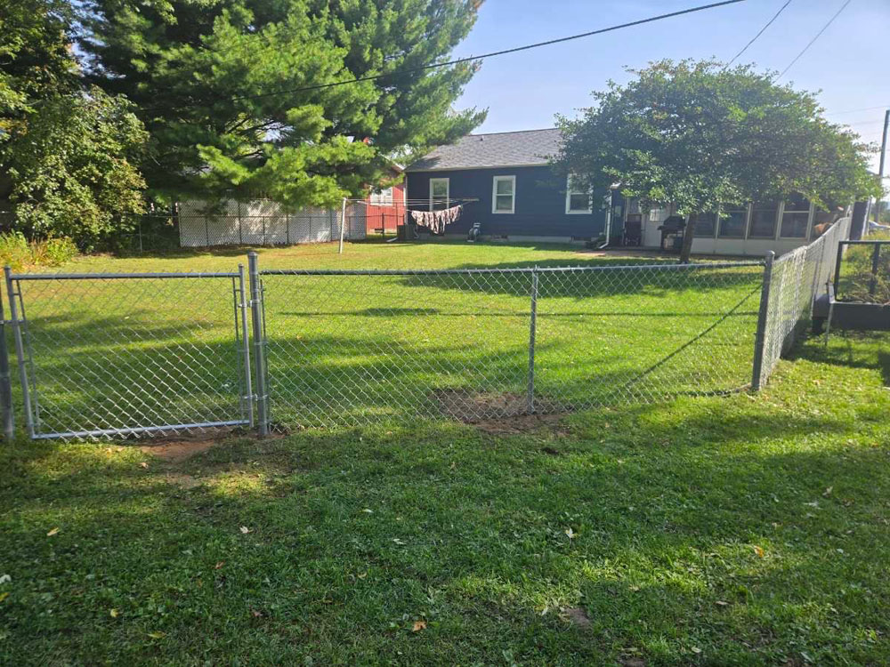 A caged metal fence in a grassy backyard, with a slightly uneven section, stands in front of a dark house with a large tree providing shade and laundry hanging on a line.