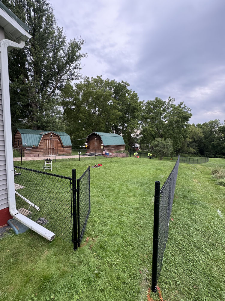 A grassy backyard is enclosed by a black chain-link fence, with several wooden sheds and tall trees in the background. Two workers in yellow vests are visible near the sheds under a cloudy sky.