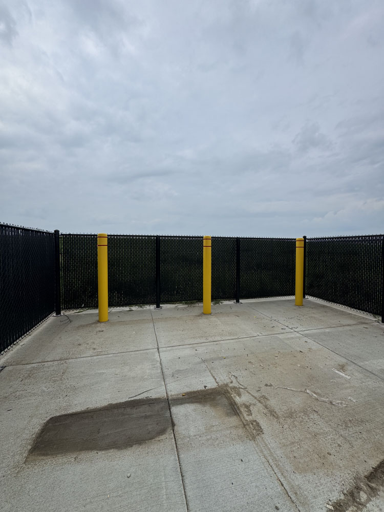 A concrete pad bordered by black chain-link fencing and four yellow bollards stands under a cloudy sky, with trees visible in the distance beyond the fence.