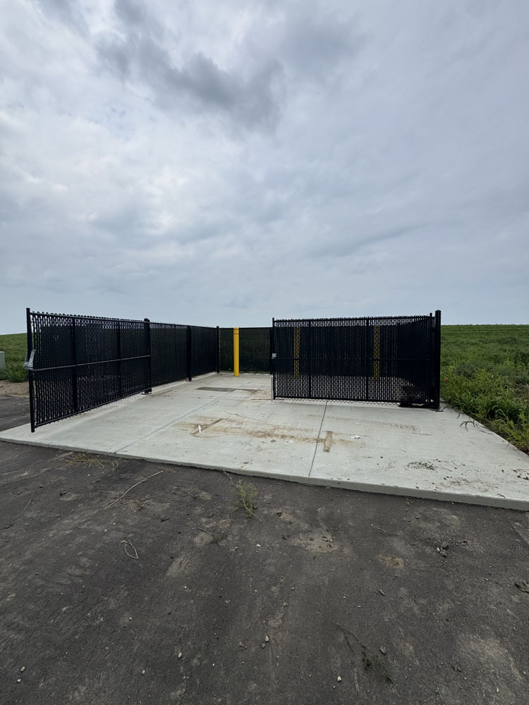 A small, empty fenced enclosure with black metal gates sits on a concrete pad under a cloudy sky, surrounded by grass and dirt. The area appears clean and unused.