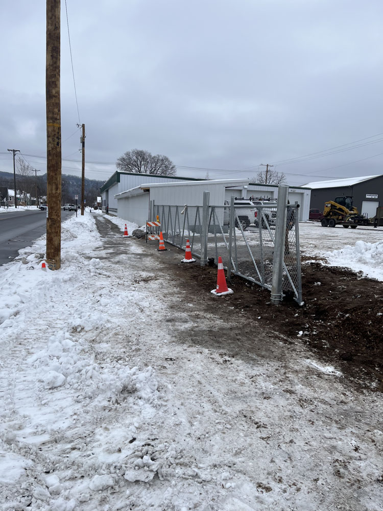 A snow-covered sidewalk runs alongside a road, with orange traffic cones placed near a chain-link fence and a utility pole. Industrial buildings and a cloudy sky are visible in the background.