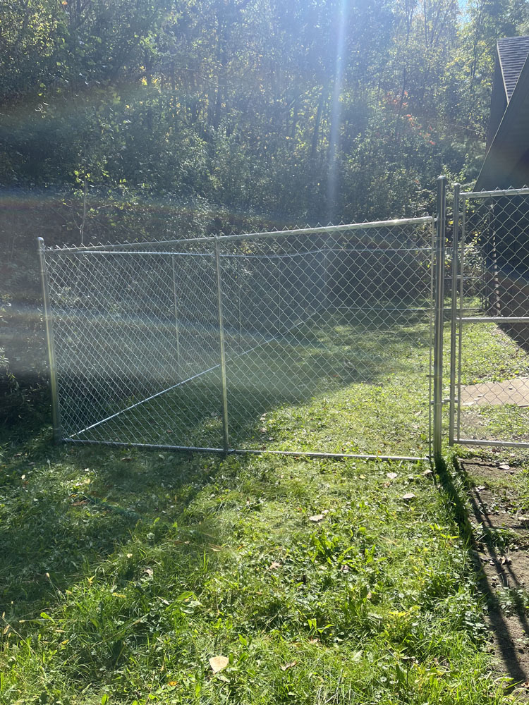 A chain-link fence section stands upright on grass, unattached at one end and not connected to any boundary, with sunlight streaming through trees in the background.