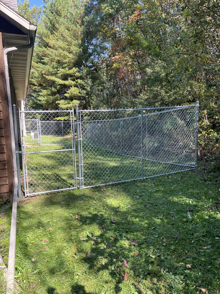 A fenced outdoor enclosure with a gate is attached to the side of a building, surrounded by green grass, trees, and shrubs under a clear blue sky.