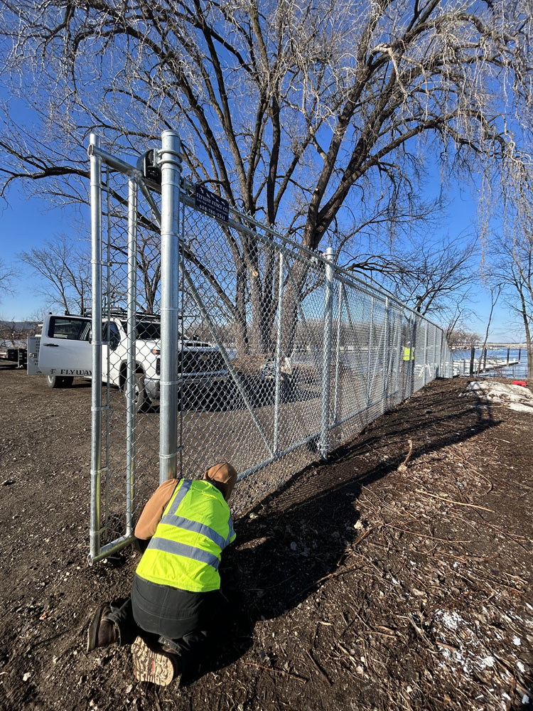 A worker in a yellow safety vest kneels by a chain-link fence, installing or inspecting it near a large leafless tree on a sunny day. Several vehicles and another worker are visible in the background.