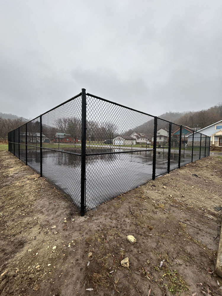 A black chain-link fence encloses a rectangular area filled with water, surrounded by bare dirt. In the background, there are houses and leafless trees under an overcast sky.