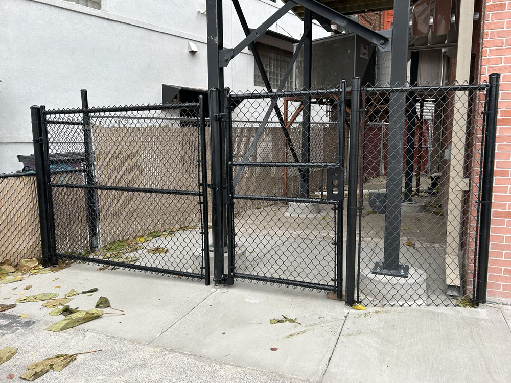 A black chain-link fence with a gate encloses an area next to a brick building. There are scattered leaves on the concrete ground, and industrial metal structures are visible inside the fenced enclosure.