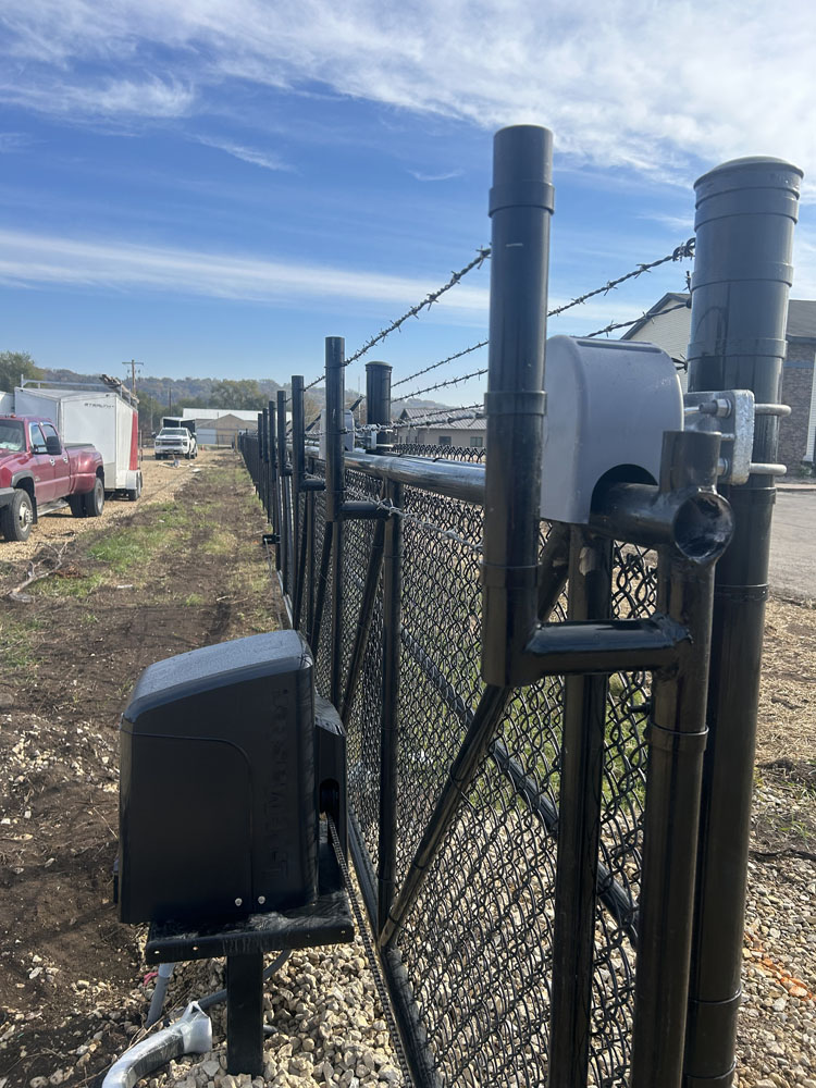 A black chain-link fence with barbed wire on top, lined with various electronic devices and utility boxes, stretches into the distance beside a dirt and gravel path. Red and white vehicles are parked on the left.