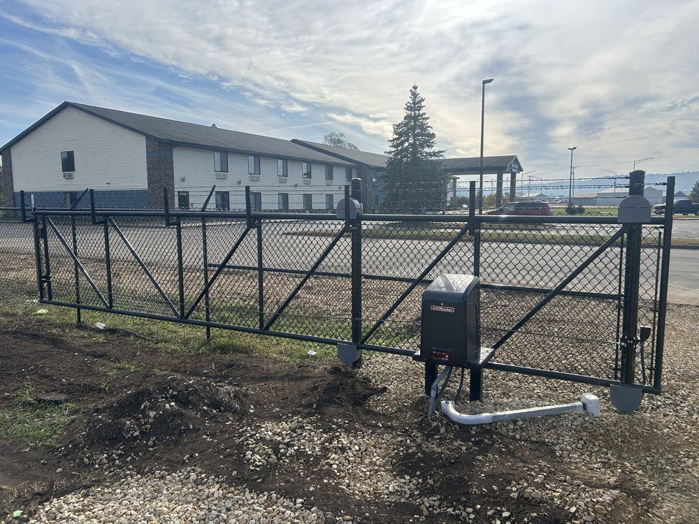 A black metal chain-link sliding gate with an automatic opener is installed at the edge of a gravel area near a road, with a building and trees visible in the background under a partly cloudy sky.