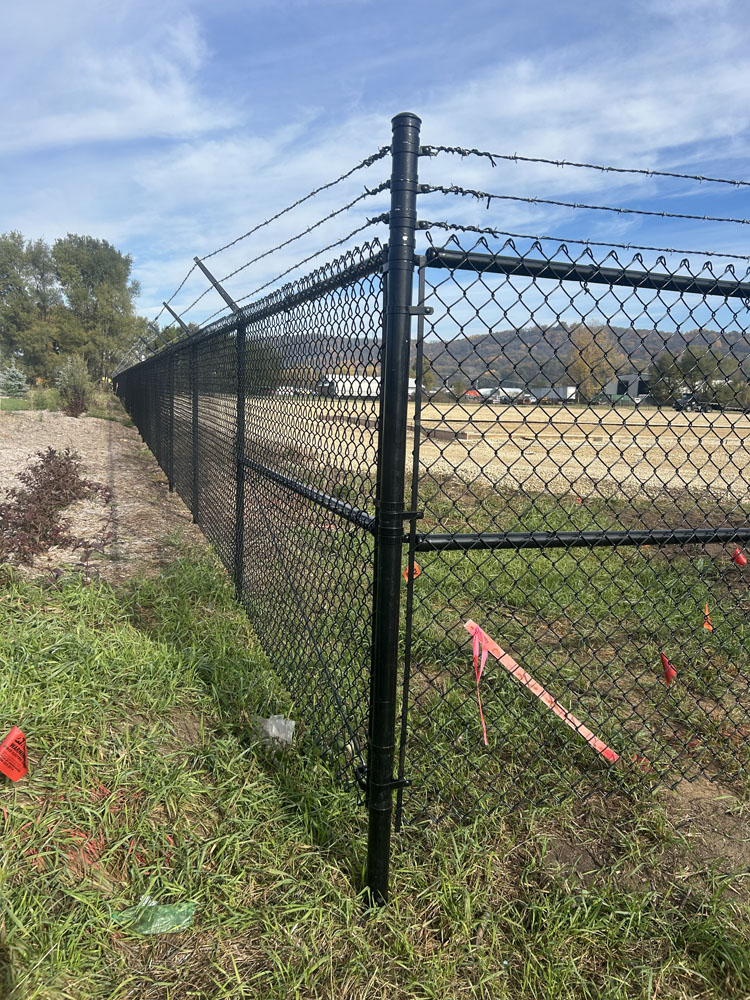 A black chain-link fence topped with barbed wire runs through a grassy area, with red marking flags on the ground and an open field in the background under a blue sky.