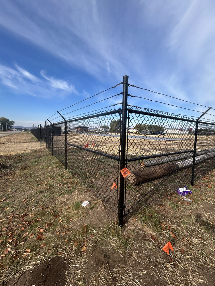 A black chain-link fence topped with barbed wire surrounds a grassy, open area under a blue sky. Orange markers and a fallen log are visible on the ground near the fence.