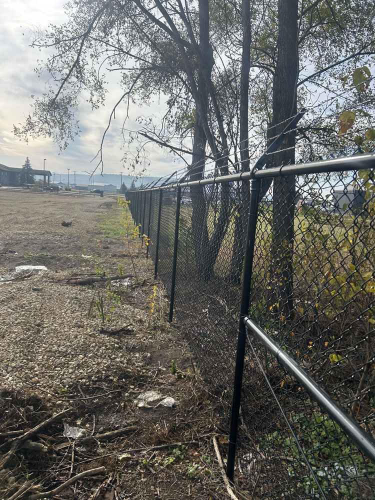 A chain-link fence topped with barbed wire runs alongside some trees, separating a gravel lot from a grassy field under a cloudy sky.