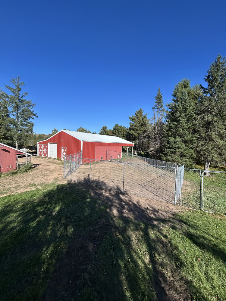 A red barn with white trim sits behind a fenced area on a sunny day, surrounded by trees and green grass under a clear blue sky.