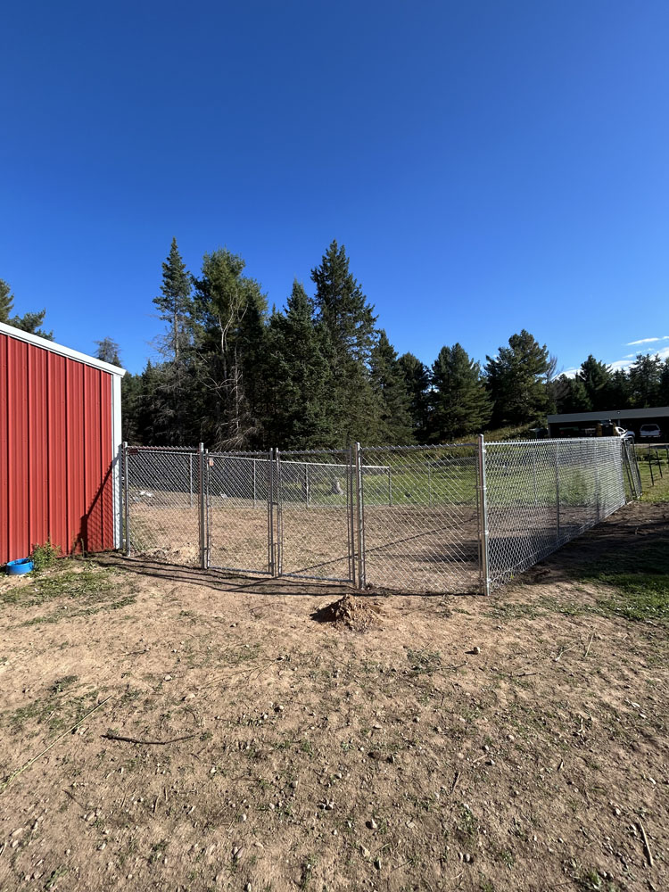 A fenced outdoor enclosure with a chain-link gate stands beside a red building, surrounded by dirt ground and green trees under a clear blue sky.