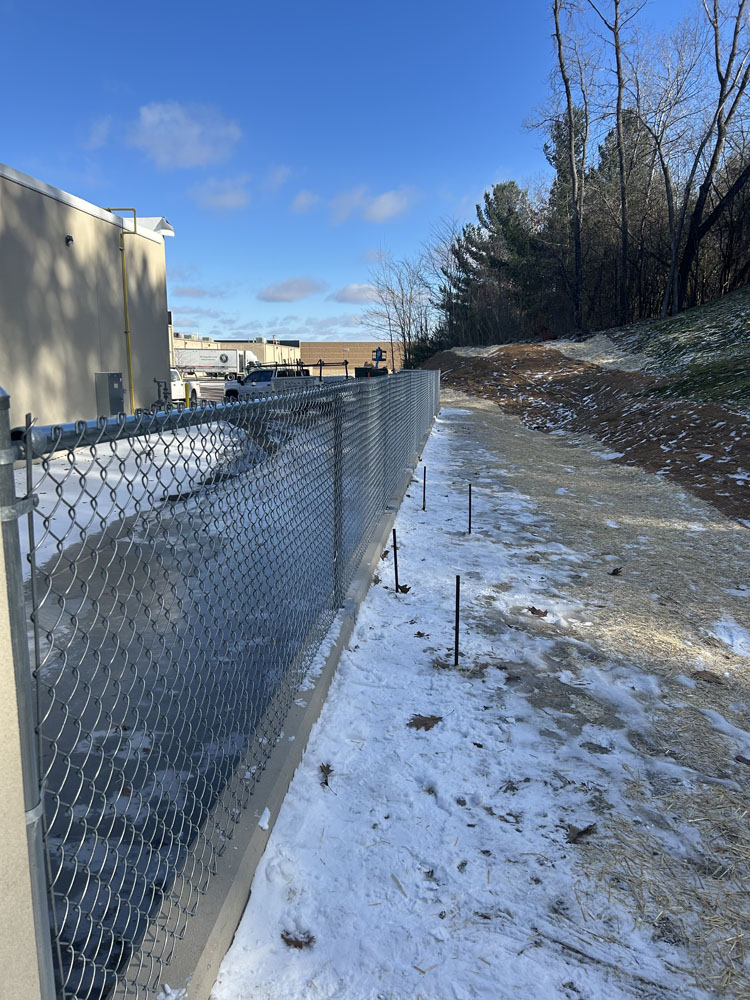 A chain-link fence runs alongside a snow-covered path beside a building, with some trees and a partially snow-dusted hill in the background under a blue sky.