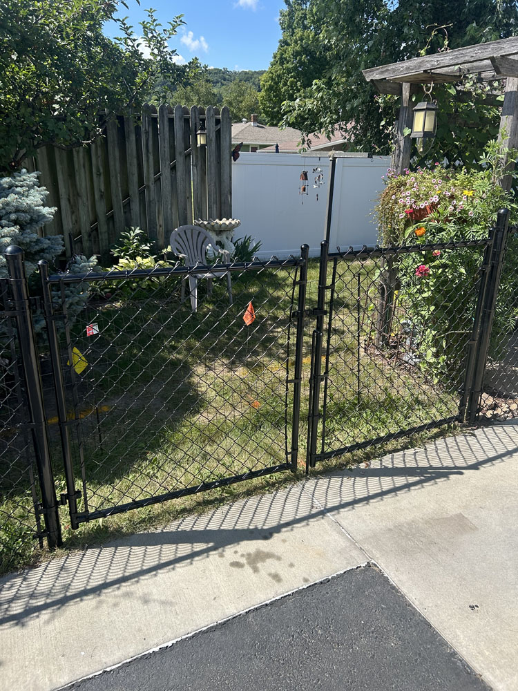 A black chain-link fence encloses a small grassy yard with a white plastic chair, garden decorations, and colorful flowers; a wooden trellis and lantern are on the right side. Shadows fall on the pavement in the foreground.