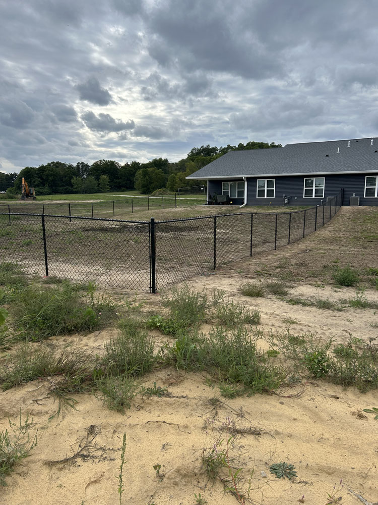 A fenced-in backyard with sparse grass and patches of sand, next to a gray house under a cloudy sky. Construction equipment is visible in the distance near a tree line.