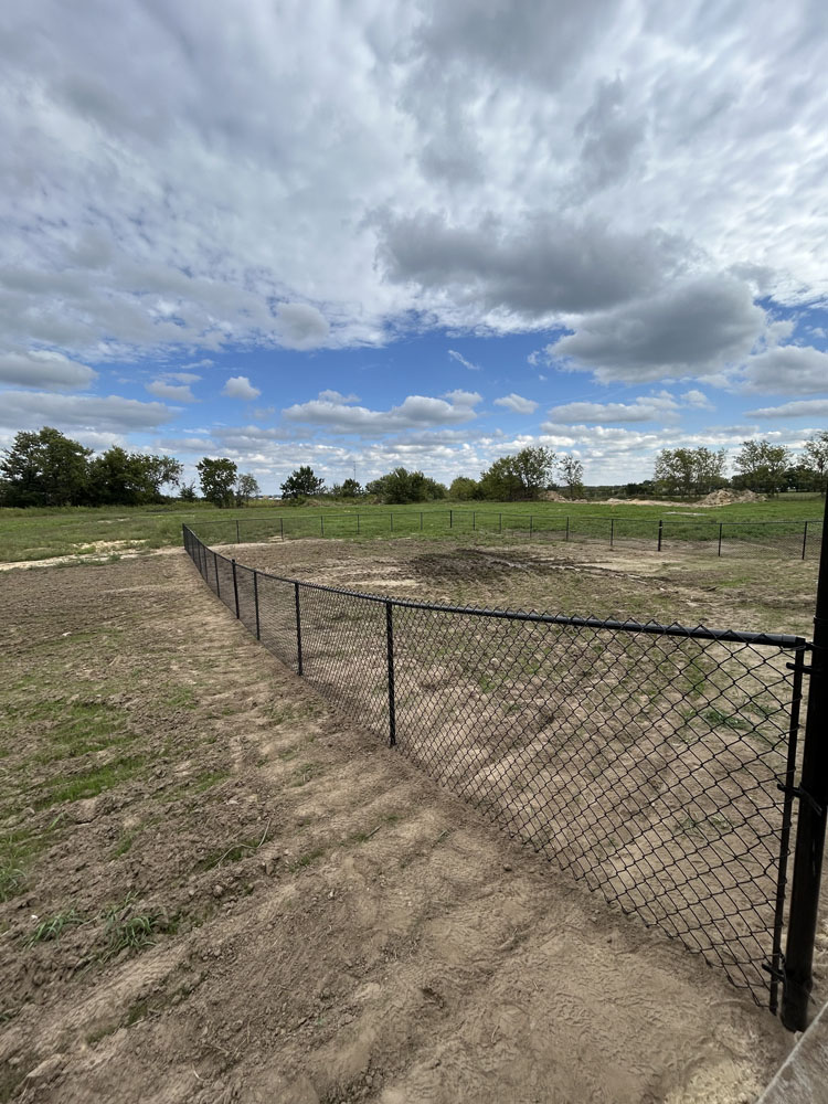 A black chain-link fence runs through a mostly barren dirt field, with patches of grass and green trees in the distance under a partly cloudy sky.