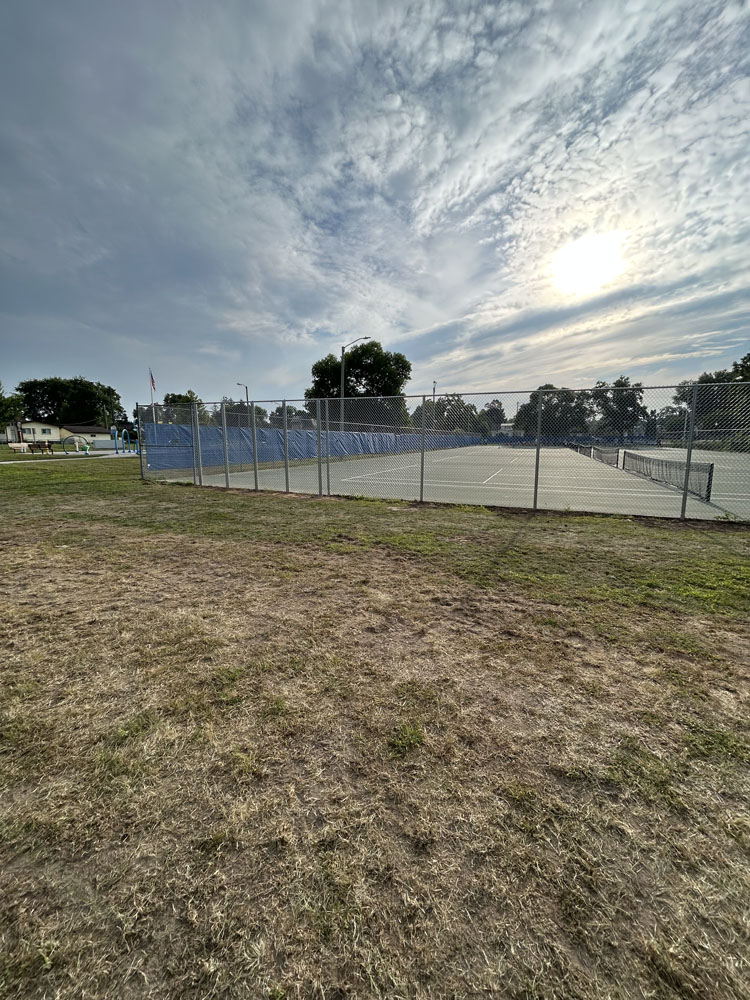 A fenced outdoor tennis court with blue wind screens sits under a partly cloudy sky as the sun shines through. The foreground shows a patchy grass field, and trees are visible in the background.