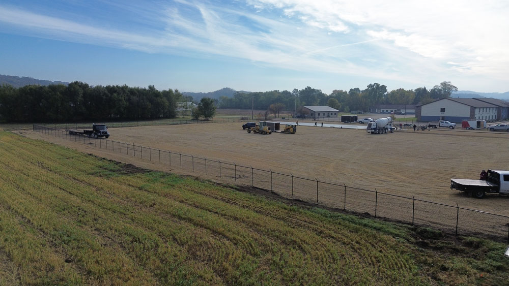 A fenced, cleared dirt lot with several trucks and construction equipment; buildings and trees are in the background under a partly cloudy sky.