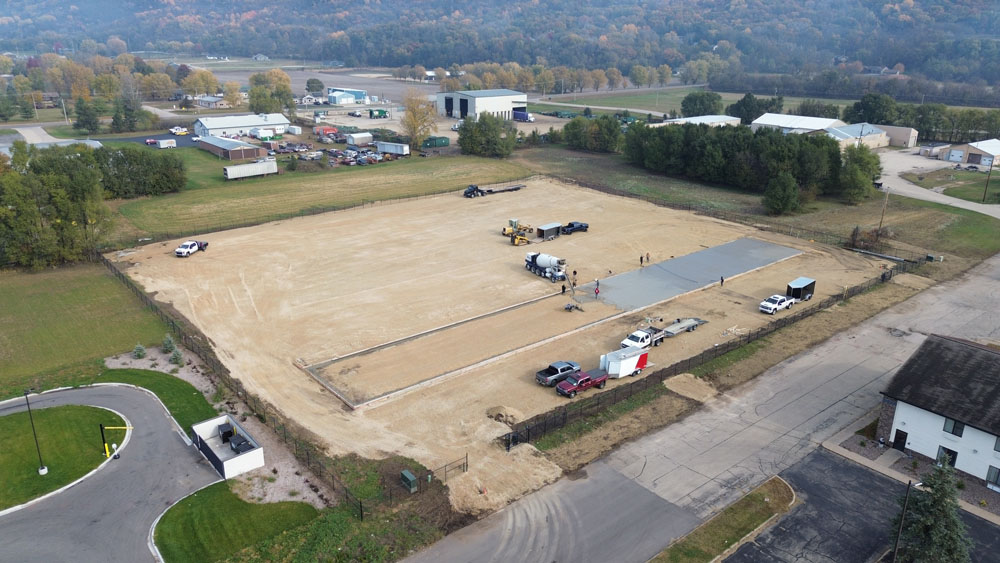 Aerial view of a construction site with vehicles and equipment on a large cleared plot of land. Workers are laying concrete slabs, surrounded by grassy fields, trees, and nearby buildings.