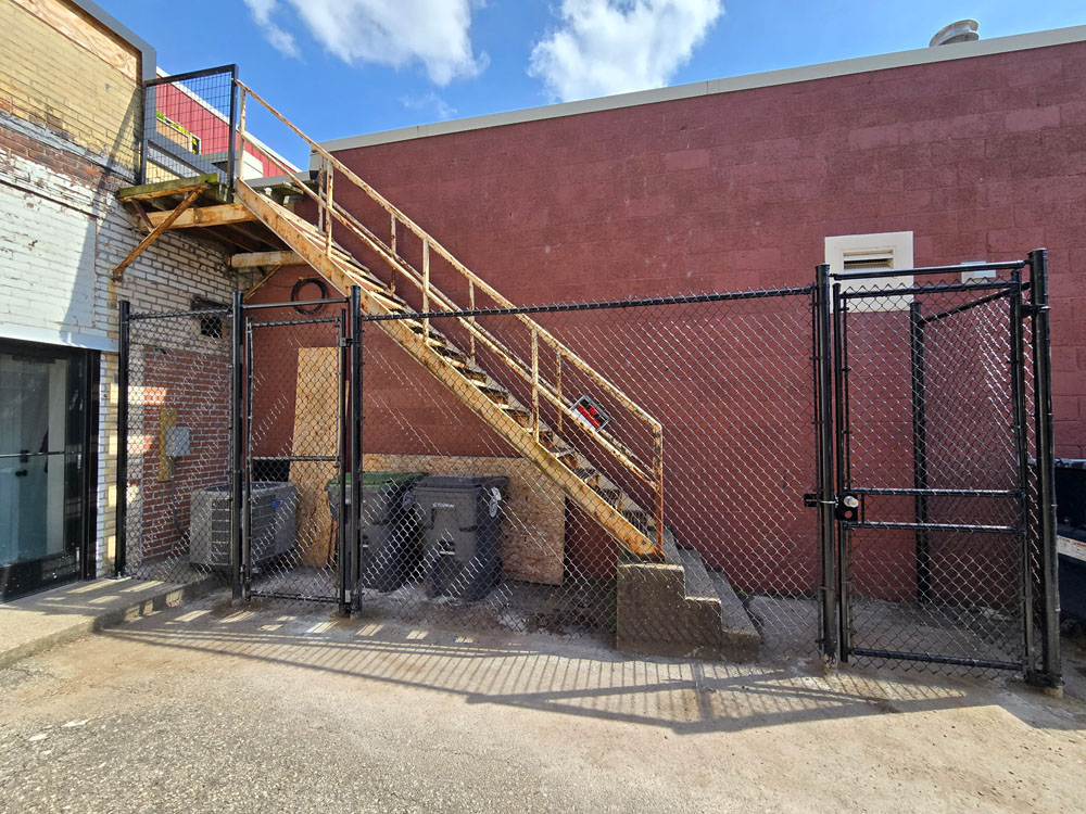 A metal staircase leads up the side of a brick building, fenced off with a black chain-link fence. Trash bins are stored underneath the stairs, and the sky is partly cloudy.