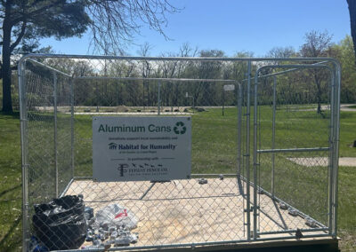 A metal fenced enclosure labeled for aluminum can recycling stands on grass in a park. Some aluminum cans and bags are scattered on the ground just outside the enclosure. Trees and open field are visible in the background.