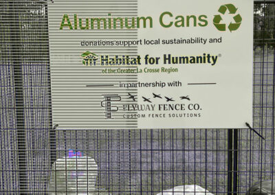 A sign on a metal cage reads "Aluminum Cans" with a recycling symbol. It notes donations support Habitat for Humanity and mentions a partnership with Beylay Fence Co. Bags of cans are visible at the bottom.